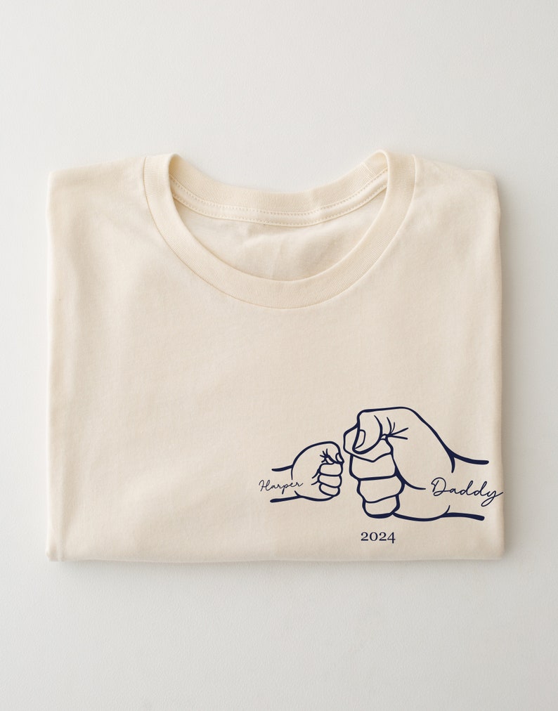 May include: A cream colored t-shirt with a graphic of two fists bumping, one labeled "Harper" and the other "Daddy". The year 2024 is printed below the graphic.