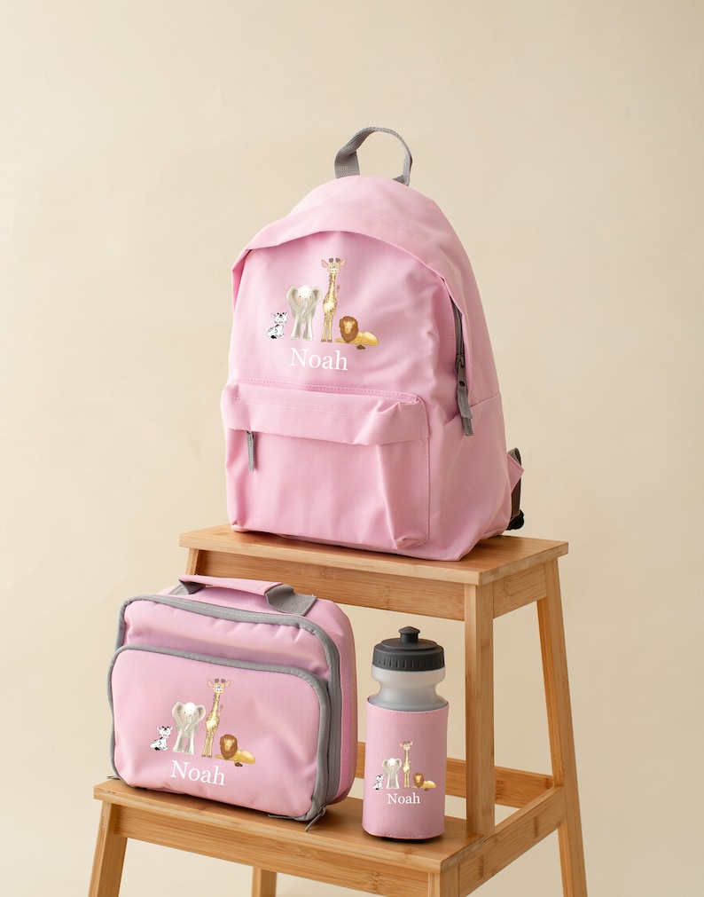May include: Pink personalized backpack, lunch bag, and water bottle set with a safari animal theme. The set features a giraffe, elephant, lion, and zebra. The name "Noah" is embroidered on each item.