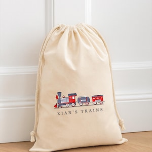 May include: A cream-coloured drawstring bag with a colourful train illustration in red, grey, and blue. The text "KIAN'S TRAINS" is printed beneath the train. The bag is made of soft fabric and has a drawstring closure.