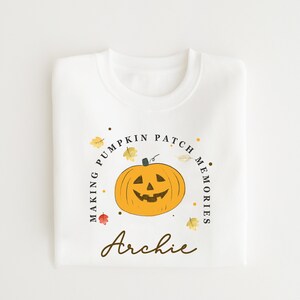 Peut inclure: Sweat-shirt blanc avec un motif de citrouille. Le motif représente une citrouille avec un visage souriant, entourée de feuilles et du texte "Making Pumpkin Patch Memories". Le nom "Archie" est écrit en cursive sous le motif.