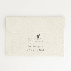 May include: A beige envelope with a black and white illustration of a stork carrying a baby in a bag. The text "Our Journey to BABY JONES" is printed below the illustration.