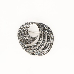 May include: A silver brooch with a swirling, openwork design. The brooch is encrusted with small, round, dark-coloured stones, creating a textured pattern. The pin is set against a white background.