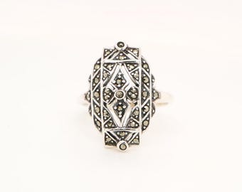 Puede incluir: Un anillo de plata con una cara ovalada y ornamentada. La cara presenta un diseño geométrico con pequeñas piedras oscuras engastadas en la plata. El anillo tiene una estética vintage o antigua, adecuado para colecciones de joyas.