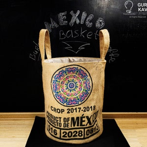 Könnte beinhalten: Ein zylindrischer Korb aus Jute mit Griffen, der ein farbenfrohes kreisförmiges Design aufweist. Auf dem Hintergrund steht der Text "MEXICO BASKET". Der Boden des Korbs trägt die Aufschrift "PRODUCT OF MÉX" und "CROP 2017-2018".