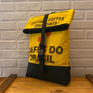 Könnte beinhalten: Ein gelber Rolltop-Rucksack mit schwarzem Text, der SPECIAL COFFEE COOPERVASS und CAFES DO BRASIL lautet. Die Tasche hat einen dunkelgrünen vertikalen Riemen mit Kunststoffschnalle und einen dunkelgrünen Boden aus einem strukturierten Material.