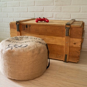 May include: A wooden crate with a red video game controller on top. The crate is sitting on a light wood floor. A brown burlap ottoman is in the foreground.