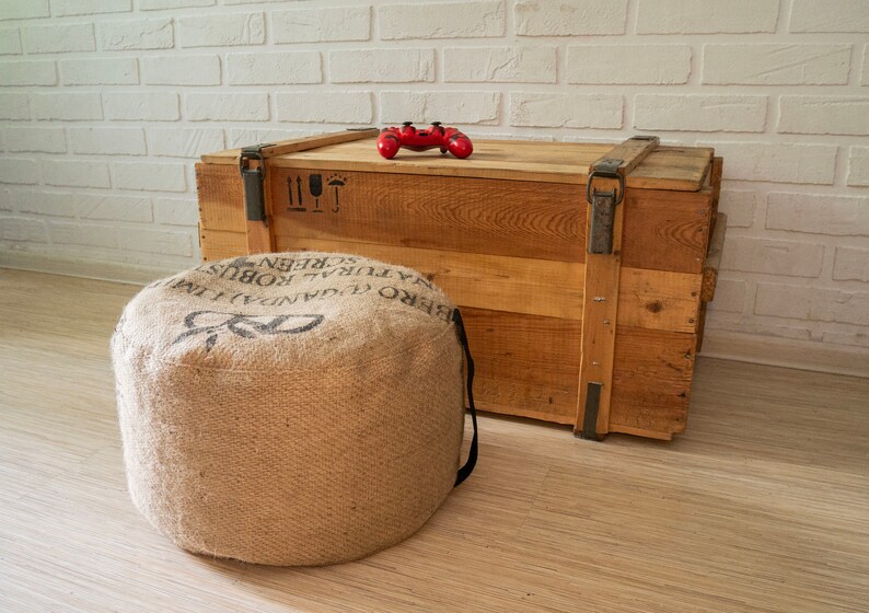 May include: A wooden crate with a red video game controller on top. The crate is sitting on a light wood floor. A brown burlap ottoman is in the foreground.