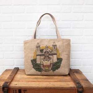 Pode incluir: Uma bolsa de lona marrom com um desenho branco de uma mulher segurando uma tigela na frente de um prédio com telhado amarelo. A bolsa tem alças de couro marrom.