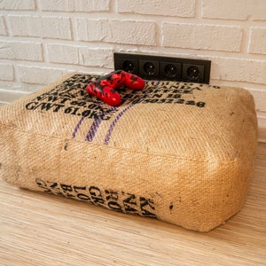 Peut inclure: Un pouf carré en toile de jute avec du texte noir et des rayures violettes. Une manette rouge et noire est posée dessus. Le pouf est contre un mur de briques blanches et un sol en bois.