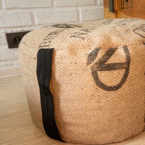 May include: A round, brown, burlap ottoman with black straps. The ottoman has a woven texture and is decorated with a black graphic.