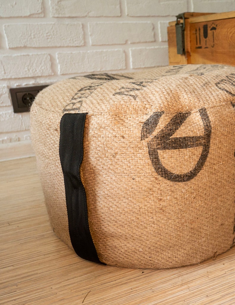 May include: A round, brown, burlap ottoman with black straps. The ottoman has a woven texture and is decorated with a black graphic.