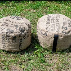 May include: Two brown burlap bean bags with black straps. The bean bags are labeled with black text that reads "UGANDA 35/119/24 60KG NET WEIGHT KG GROSS WEIGHT FOR EXPORT ONLY" and "(UGANDA) LIMITED NATURAL ROBUST SCREEN CROP COFFEE PRODUCE UG 35/119/24".