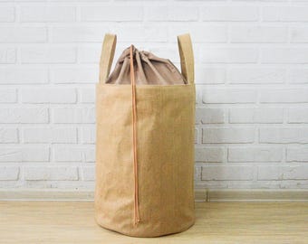 May include: A cylindrical, tan storage basket with two handles. The basket has a drawstring closure and a light brown interior. The exterior is made of a textured fabric, and a thin, orange-brown cord runs down the front.