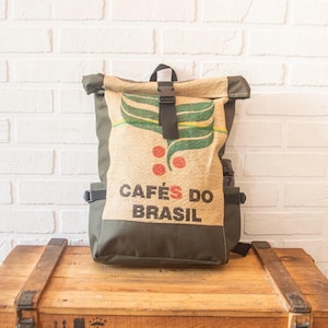 Może przedstawiać: Plecak z motywem worka na kawę. Z przodu napis "CAFÉS DO BRASIL" w kolorze czarnym, z ilustracją zielonego liścia i czerwonych jagód. Posiada rolowane zamknięcie z czarną klamrą i oliwkowozielone boki.