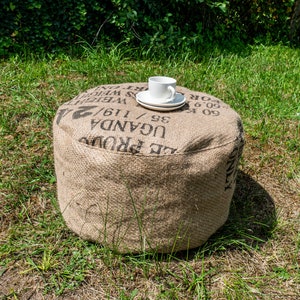 May include: A round, brown, burlap ottoman with black text that reads "UGANDA 35/116/ WEIGHT 2", a white ceramic cup and saucer on top.
