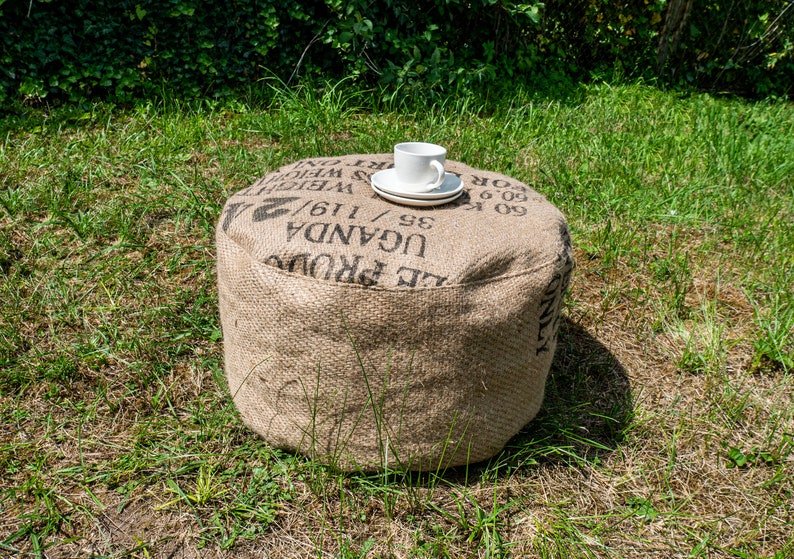May include: A round, brown, burlap ottoman with black text that reads "UGANDA 35/116/ WEIGHT 2", a white ceramic cup and saucer on top.