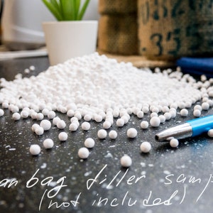 May include: A close-up of white bean bag filler beads scattered on a black surface. A blue pen is lying on the surface. The text "Bean bag filler samples (not included)" is written on the surface.