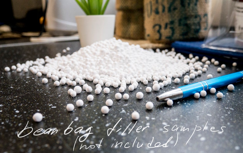 May include: A close-up of white bean bag filler beads scattered on a black surface. A blue pen is lying on the surface. The text "Bean bag filler samples (not included)" is written on the surface.