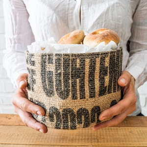 Könnte beinhalten: Eine Person hält einen Jutesack mit dem Aufdruck "Garten". Der Sack ist mit Brötchen gefüllt.