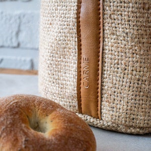 Könnte beinhalten: Ein brauner Jutespeicherbehälter mit einem Lederriemen und dem Wort "CARME" geprägt auf dem Riemen. Der Behälter steht auf einer weißen Oberfläche mit zwei braunen Baguettes im Vordergrund.