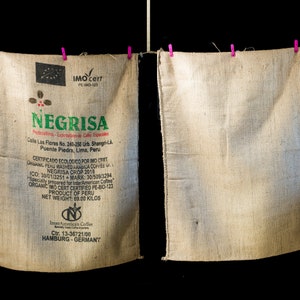 Può includere: Due sacchi di juta appesi a uno stendino. Il sacco a sinistra ha un'etichetta con il testo "NEGRISA" e informazioni sul prodotto, tra cui "CERTIFICADO ECOLOGICO POR IMO CERT" e "PRODUCT OF PERU". Il sacco a destra è vuoto.