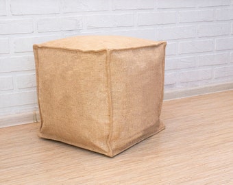 May include: A square-shaped ottoman covered in natural burlap fabric. The ottoman has a simple, rustic design and is perfect for adding a touch of texture to any room.