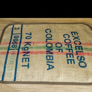 May include: A brown burlap coffee bag rug with blue text that reads "EXCELSO COFFEE OF COLOMBIA 70 KG NET 3 10068".