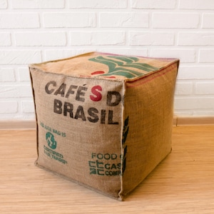 Pode incluir: Um pufe quadrado de juta marrom com as palavras "Café's do Brasil" impressas na frente. O pufe tem um design verde e branco na parte superior.