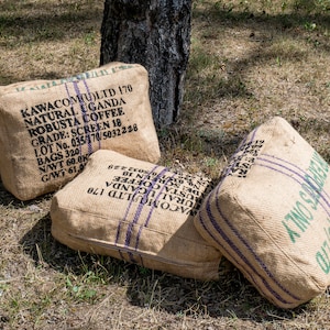 May include: Three burlap sacks with black and white text and purple stripes. The text on the sacks reads "KAWACOM(U)LTD 170 NATURAL UGANDA ROBUSTA COFFEE GRADE: SCREEN 18 LOT No. 035/170/5032228 BAGS 320 N/WT 60.0K G/WT 61" and "REIPOLES ONLY".