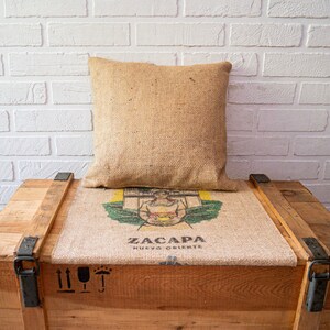 May include: A wooden crate with a burlap cushion and a pillow. The cushion has a printed image of a woman holding a cup and the text "ZACAPA NUEVO ORIENTE".
