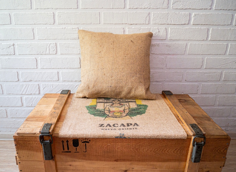 May include: A wooden crate with a burlap cushion and a pillow. The cushion has a printed image of a woman holding a cup and the text "ZACAPA NUEVO ORIENTE".