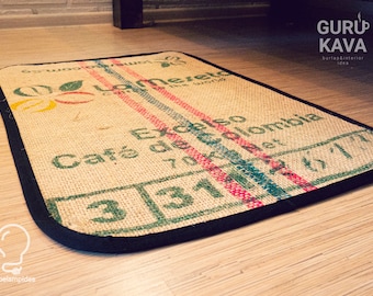 May include: A brown burlap rug with a black border. The rug has a green, red, and blue stripe pattern and the text "Cafe de Colombia" and "Excelso" printed on it.