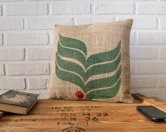 May include: A brown burlap pillow with a green leaf design and a red dot in the center. The pillow is sitting on a wooden surface with the text "HE BROGATH!" and some numbers.