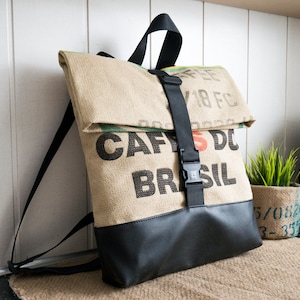May include: A brown canvas backpack with black straps and a black leather bottom. The front of the backpack is made from a repurposed coffee bag with the text "CAFES DO BRASIL" printed on it.