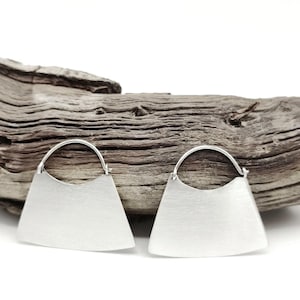 May include: A pair of silver-toned earrings with a unique, geometric design. Each earring features a flat, trapezoid-shaped body with a curved top and a circular hoop for the ear. The earrings are displayed against a backdrop of weathered wood.