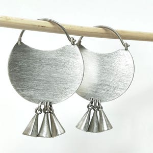 May include: A pair of silver-toned earrings. Each earring features a large, brushed metal, crescent-shaped disc with three small, cone-shaped dangles. The earrings are suspended from a silver wire. The earrings are displayed on a wooden dowel.