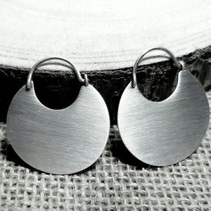 May include: A pair of silver-toned, circular earrings with a brushed metal finish. Each earring features a crescent-shaped cutout at the bottom and a curved hook at the top for attachment. The earrings are displayed on a textured, neutral-toned surface.
