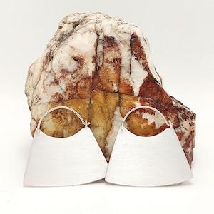 May include: A pair of silver-toned earrings featuring a wide, fan-shaped metal plate with a brushed finish and a thin, curved wire hook at the top. The earrings are positioned in front of a large, textured rock with white, tan, and reddish-brown tones.