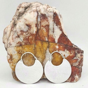 May include: A pair of silver-toned hoop earrings featuring a flat, circular disc design with a brushed metal finish. The earrings have a simple wire closure mechanism and are positioned in front of a large, multi-colored rock with white, yellow, and reddish-brown mineral veins.