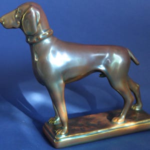 Puede incluir: Una figura de perro de color bronce en posición de pie. La escultura presenta un acabado liso y brillante y un collar detallado. El perro está colocado sobre una base rectangular, con una ligera variación de color.