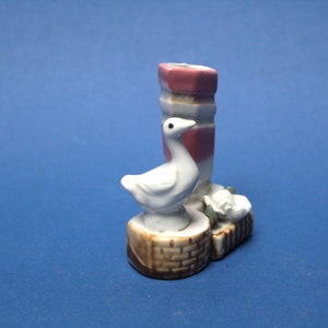 Może przedstawiać: Biała ceramiczna figurka gęsi osadzona na brązowo-białej podstawie, obok różowo-białej, pasiastych ceramicznych wazonów z białym kwiatem.