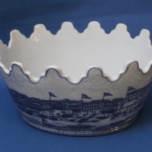 Peut inclure: Bol en céramique blanc avec un bord festonné et un motif de paysage urbain bleu et blanc à l'extérieur. Le bol a une large ouverture et une illustration détaillée de bâtiments et de drapeaux.