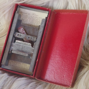 May include: A red box with a metal razor blade sharpener inside. The box is open and the sharpener is visible. The sharpener has a metal blade and a metal handle. The box is made of cardboard and has a red lining.