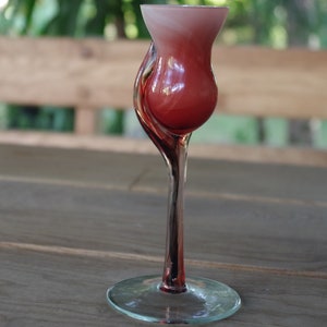 Op de afbeelding: Een hoog, helder glas met een rood en wit swirl-ontwerp. Het glas heeft een unieke, gebogen steel en een brede, ronde basis.