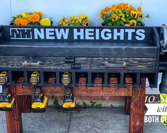 May include: A black wooden wall-mounted tool organizer with a drawer and 10 slots for storing tools. The organizer has a sign that says "NH NEW HEIGHTS" and features a wrench-shaped handle on the drawer. The organizer is mounted on a wooden table with a red stain. There are several yellow and black power drills hanging from the organizer. A green and black circular saw is mounted on the right side of the organizer. The text "10 Slots with BOTH CUSTOM" is visible in the bottom right corner of the image.