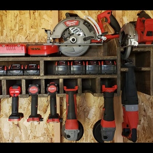May include: A wooden wall-mounted storage rack for power tools and batteries. The rack has multiple compartments for storing various tools, including drills, saws, and grinders. The rack is painted a dark brown color and has a distressed finish. The tools are all red and black and are arranged neatly on the rack.