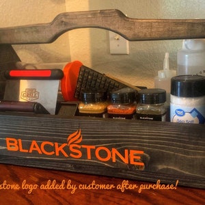 May include: A wooden tool caddy with a black finish and a handle. The caddy is filled with various grilling tools, including a spatula, a grill brush, and a bottle of sea salt. The caddy has the words "BLACKSTONE" printed on the front in orange.