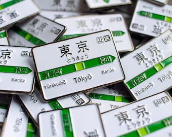 May include: A close-up of several enamel pins featuring the Tokyo Metro train line. The pins are white with green and black text and graphics. The text on the pins reads "Tokyo", "Yurakucho", "Kanda", and "とうきょう".