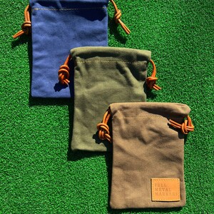 May include: Three small drawstring bags made of canvas fabric. The bags are in blue, green, and brown. The bags have leather drawstrings and are stacked on top of each other. The bottom bag has a brown leather patch with the text "FULL METAL MARKERS" on it.