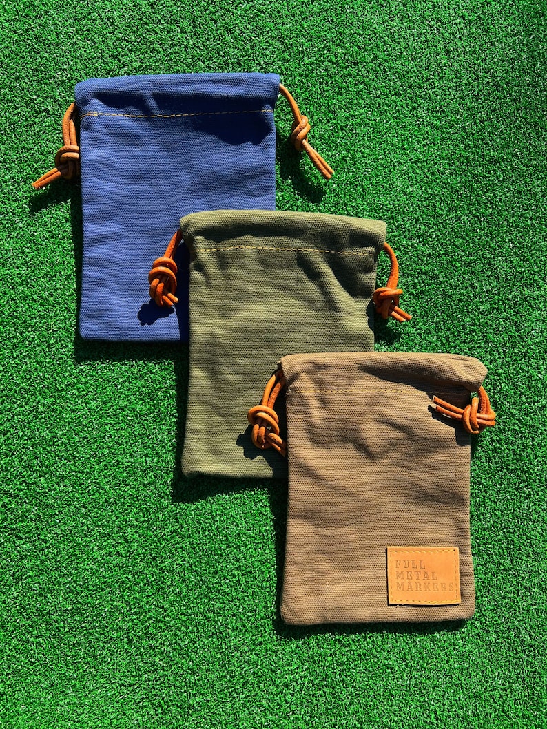 May include: Three small drawstring bags made of canvas fabric. The bags are in blue, green, and brown. The bags have leather drawstrings and are stacked on top of each other. The bottom bag has a brown leather patch with the text "FULL METAL MARKERS" on it.